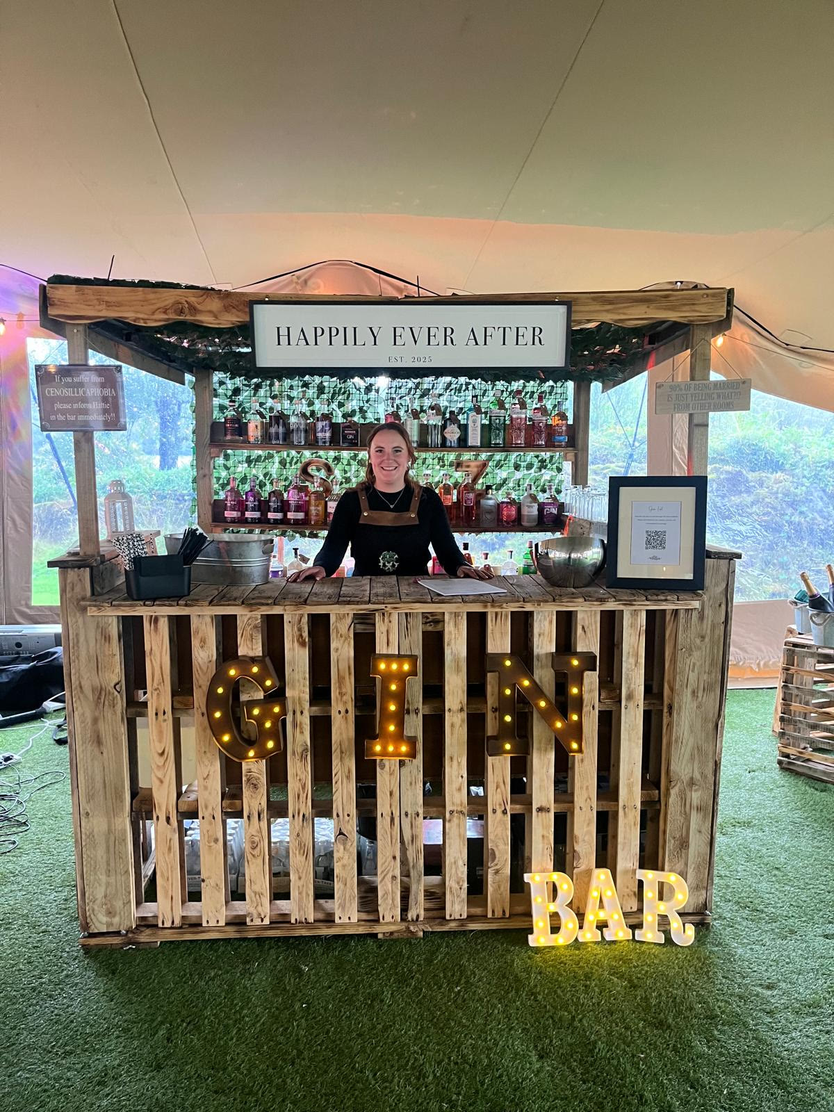 Harriet Kennedy behind the wooden Tipsy Daisy gin bar, with a Happily Ever After sign and illuminated GIN BAR letters