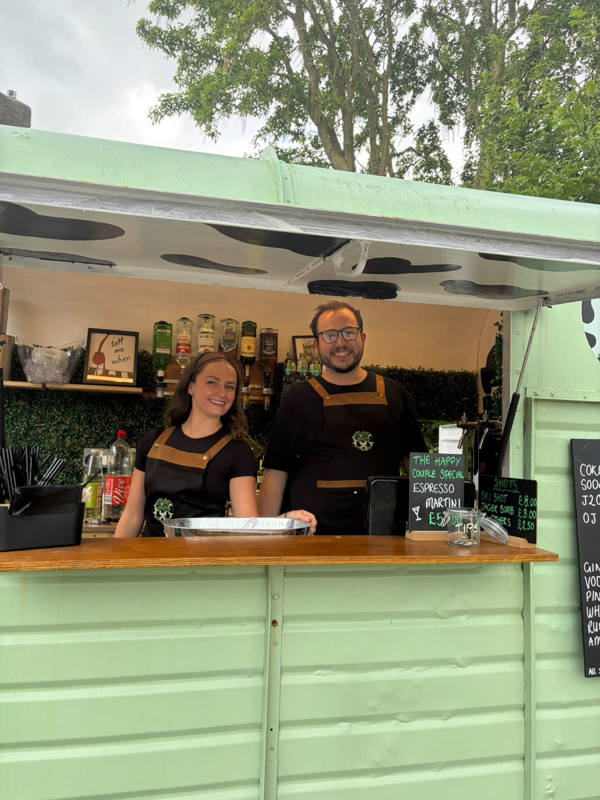 Two Tipsy Daisy bartenders smiling at the serving hatch of the sage green horsebox bar