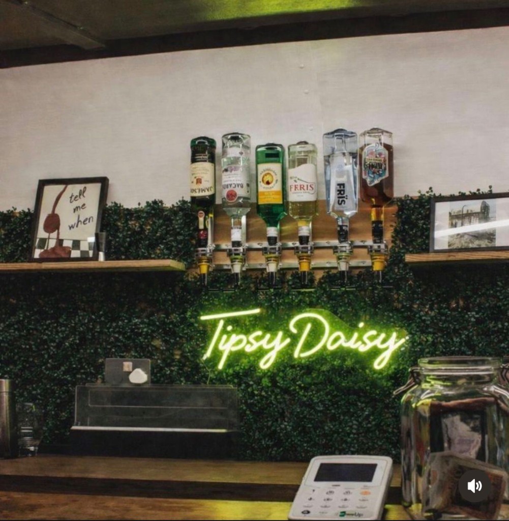 Tipsy Daisy bar interior: Tipsy Daisy neon over green foliage, back-bar optics, and wooden counter